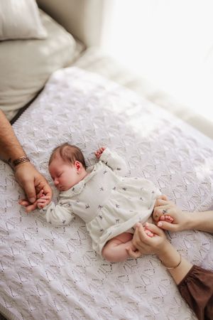 Newborn in white outfit sleeps peacefully on textured white bedding while hands reach to touch.