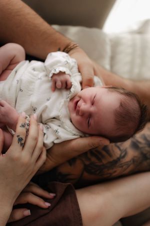 A sleeping newborn baby in white clothing rests peacefully on tattooed arms.