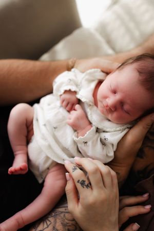 A newborn baby wrapped in white cloth being gently cradled by hands with visible tattoos.