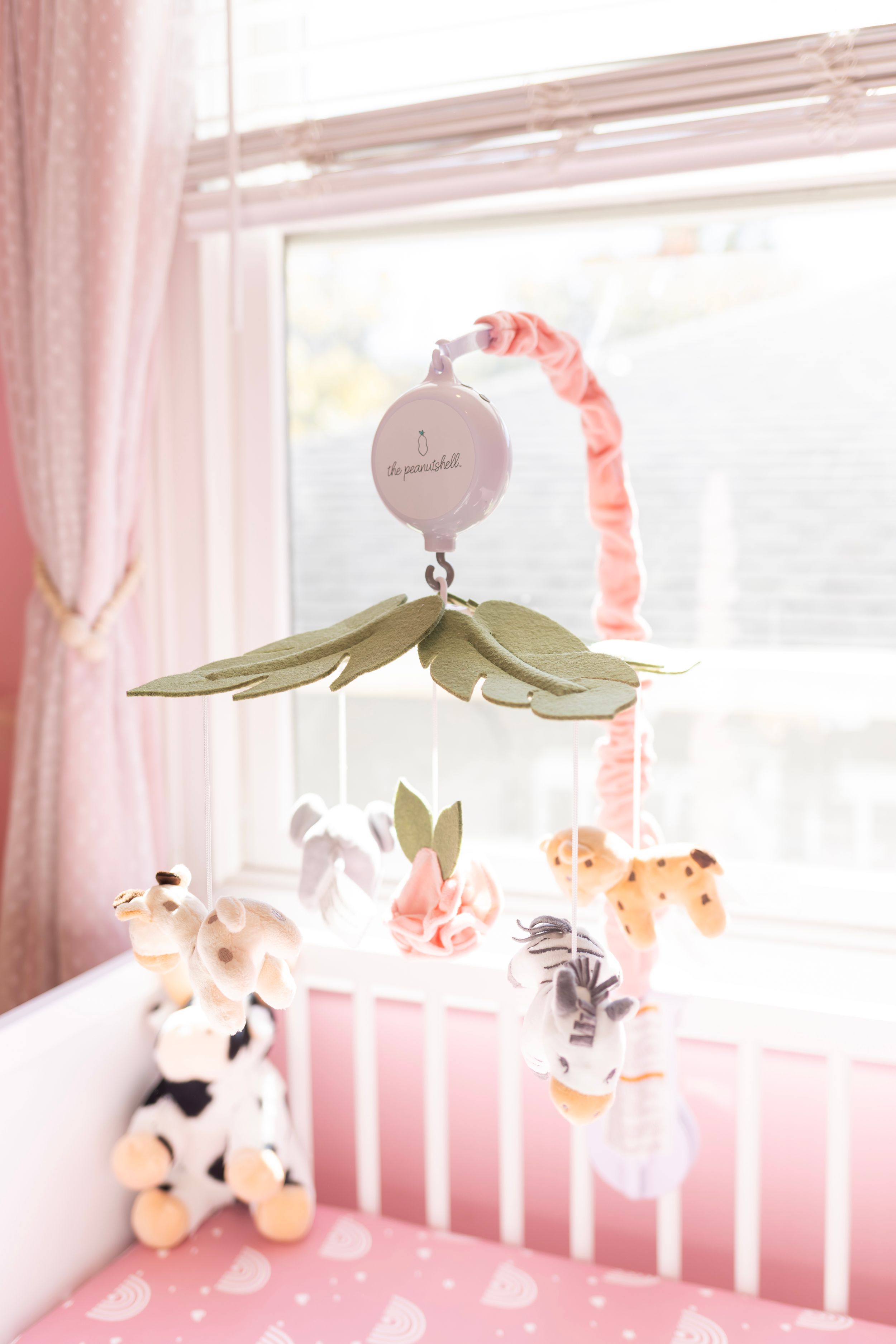 Soft plush animal toys and mobile decorations hang above a white baby crib in a nursery with pink accents.