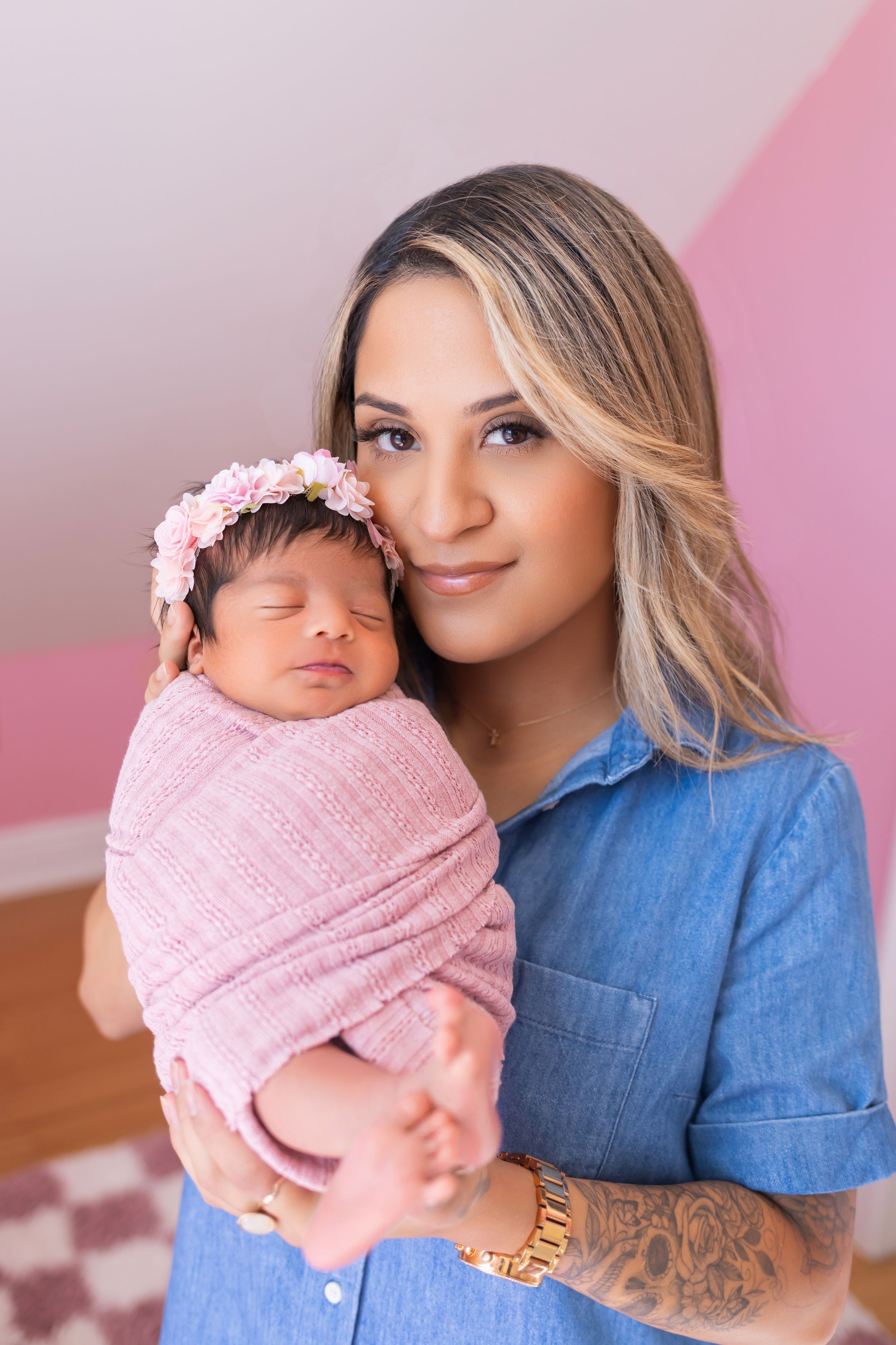 A parent in a blue denim shirt holds a sleeping newborn wrapped in a pink blanket against a pink background.
