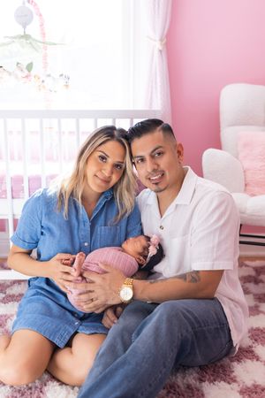 A couple sits together in a pink nursery room holding a newborn baby in a tender moment.