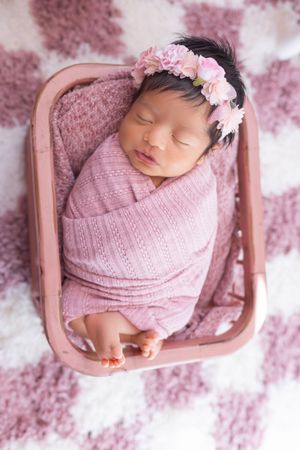 Sleeping newborn baby wrapped in pink knit blanket rests peacefully in small pink bed with delicate floral headband.