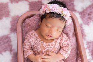 A sleeping newborn wearing pink lace and floral headband rests peacefully on a pink textured backdrop.