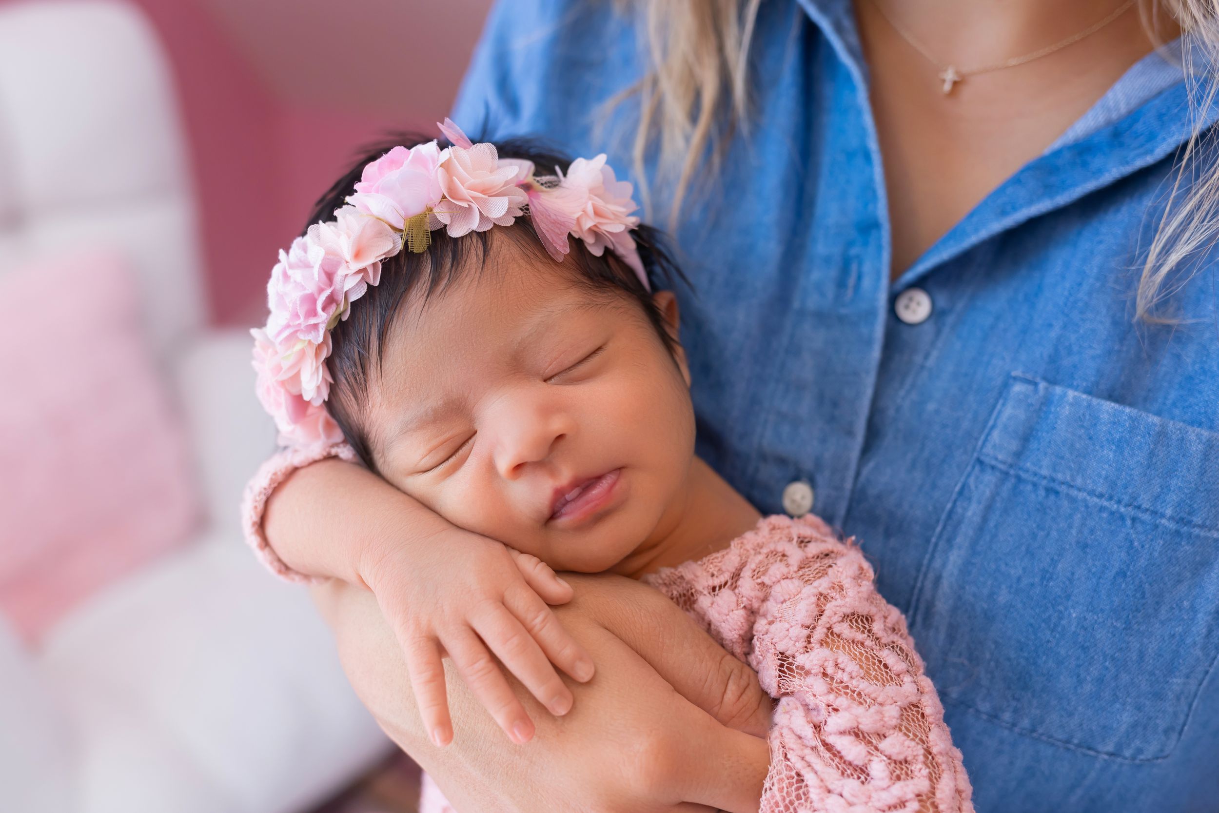 A sleeping newborn baby wearing a pink headband and floral outfit rests peacefully in a pink-toned setting.