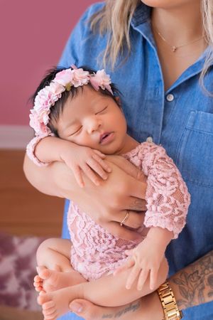 Sleeping newborn in pink lace outfit and floral headband held gently in denim-clad arms.