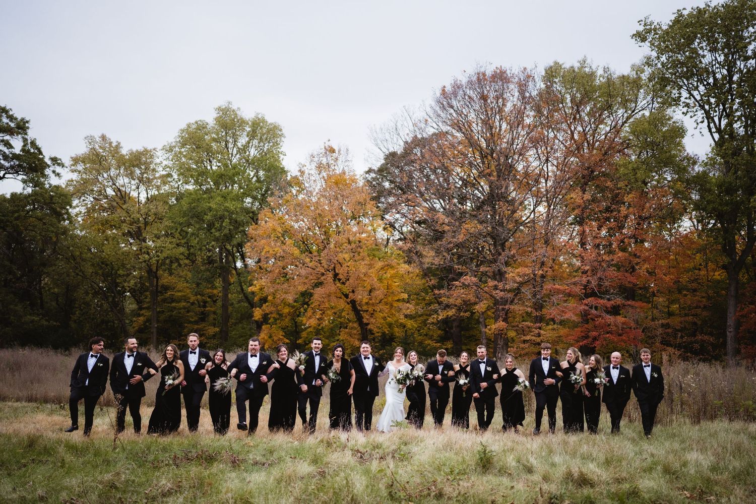 A wedding party stands in a line against autumn trees with golden foliage in a scenic outdoor setting.