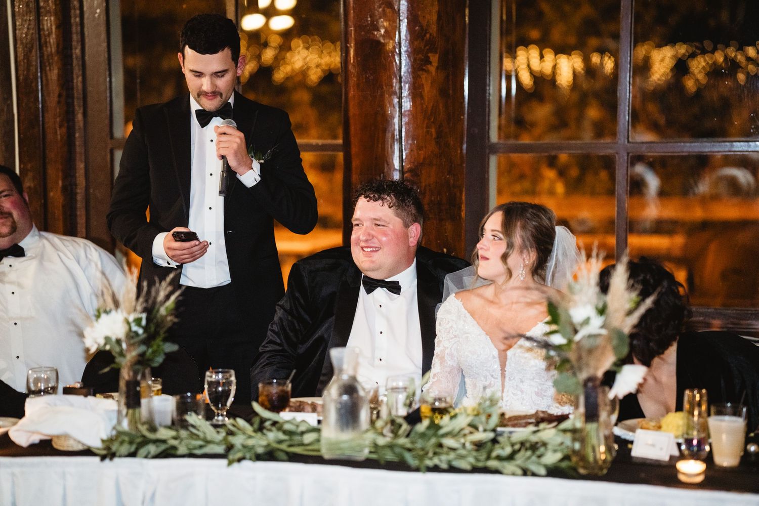 Wedding reception guests enjoy speeches and laughter at an elegant head table decorated with greenery garlands.