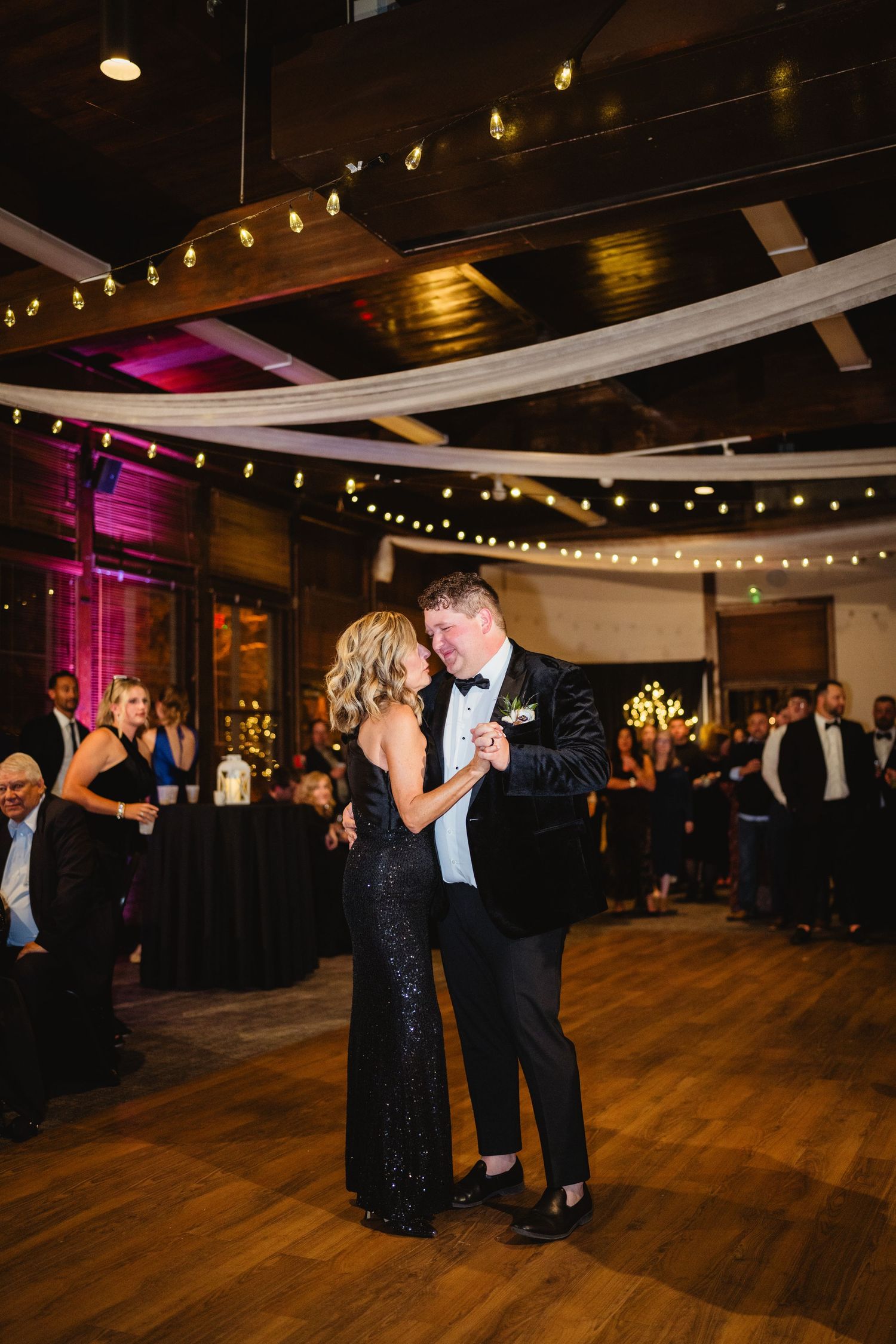 Wedding guests dance together on a dark wooden dance floor under string lights at an elegant indoor reception venue.