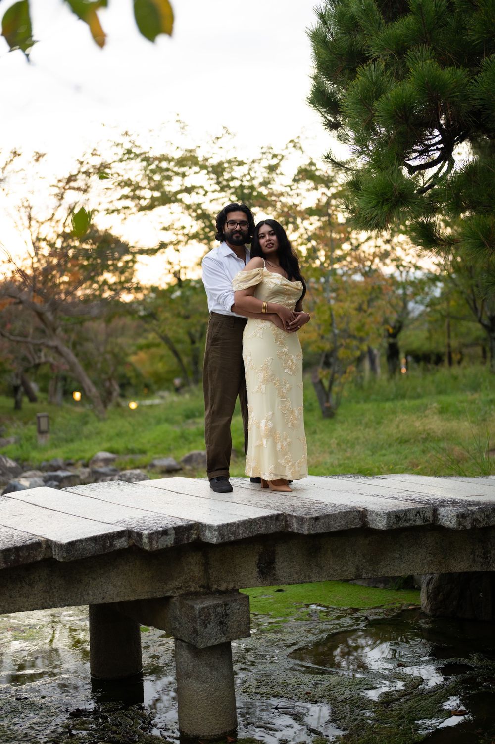 Couple embracing on a stone bridge over a pond at golden hour in a Japanese garden.