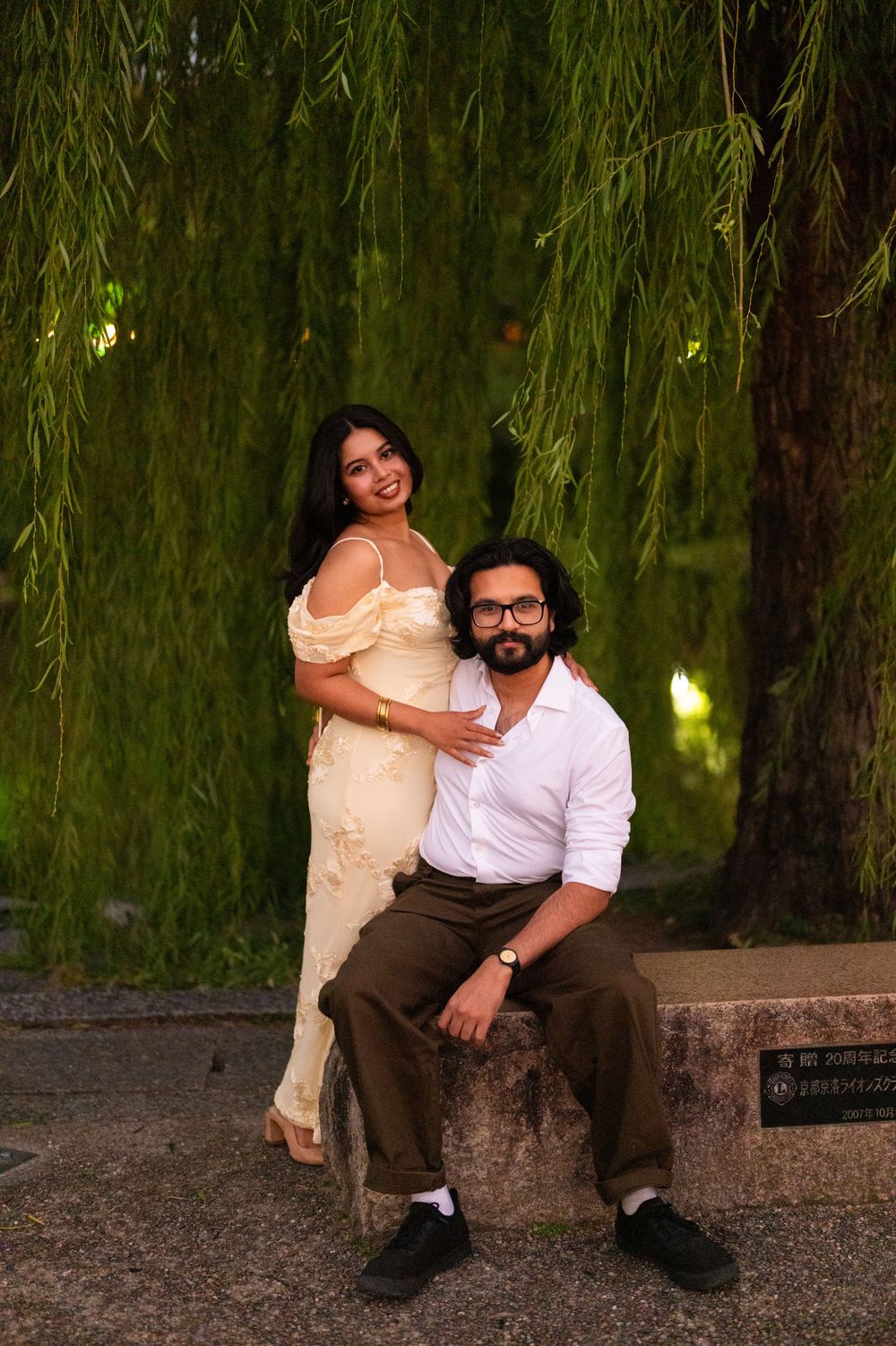 Couple posing under cascading weeping willow branches, woman in cream dress embracing seated man.