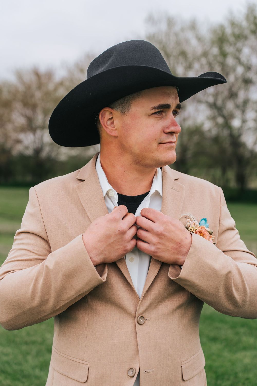 A person in a tan blazer and cowboy hat adjusts their collar while standing outdoors.