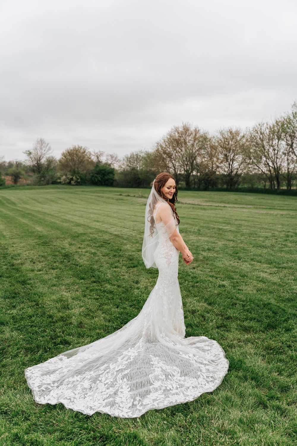 A bride in a lace mermaid wedding gown with long train poses on a green lawn under an overcast sky.