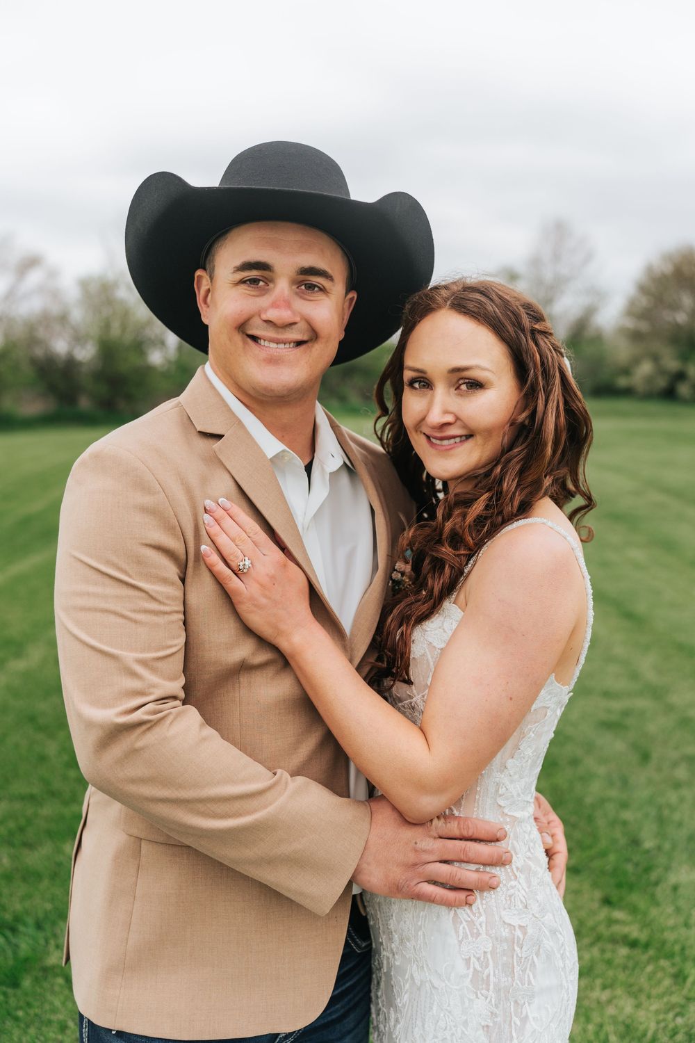 A couple in western-inspired wedding attire embraces in a green field on their special day.