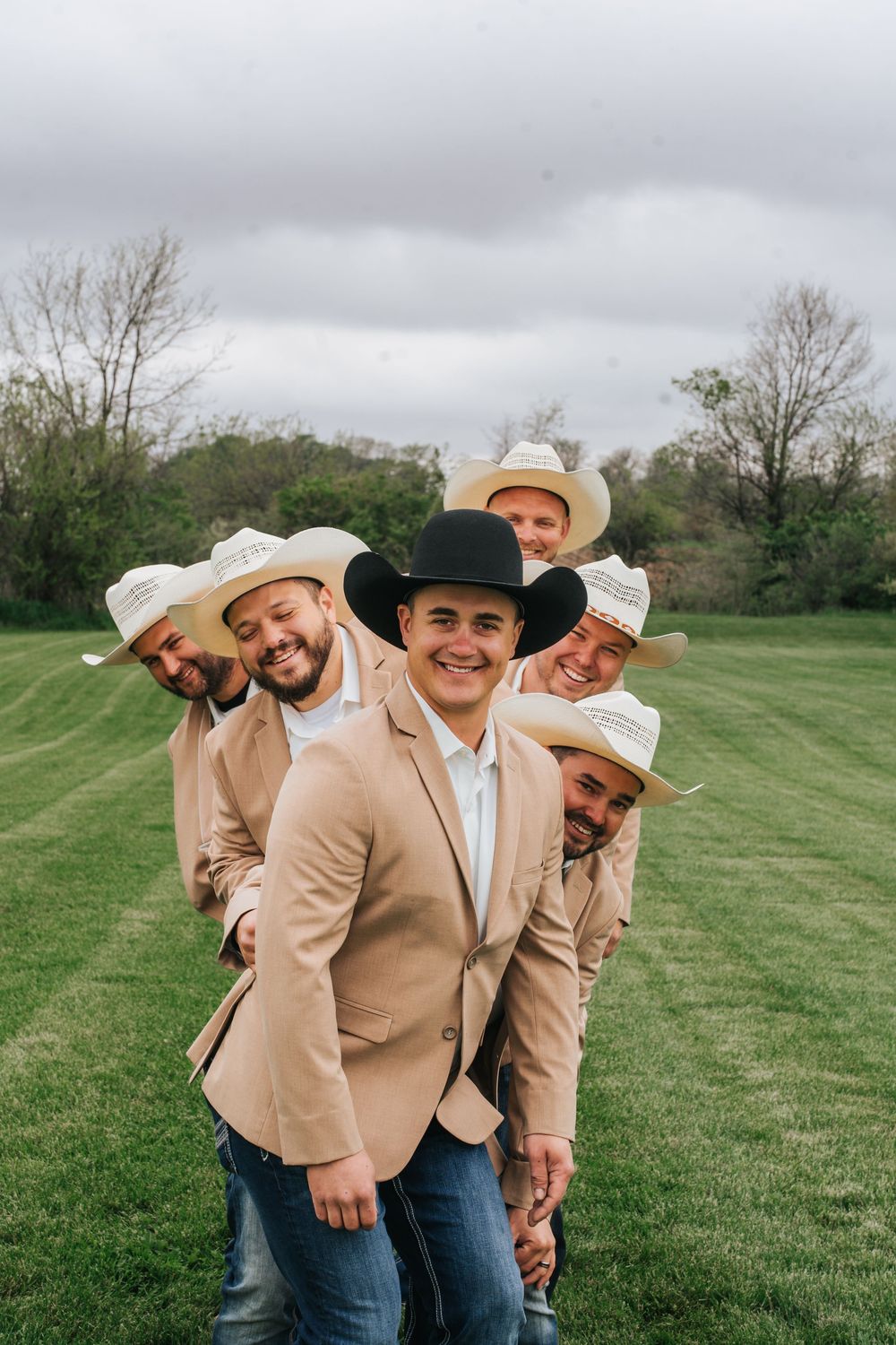 A group in matching beige jackets and cowboy hats pose playfully in a grassy field on a cloudy day.