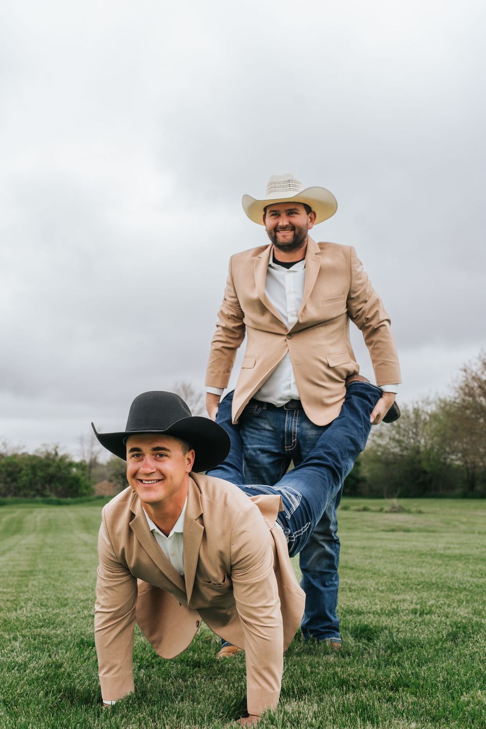 Three playful people in tan blazers and cowboy hats crawl on green grass during a fun outdoor photoshoot.