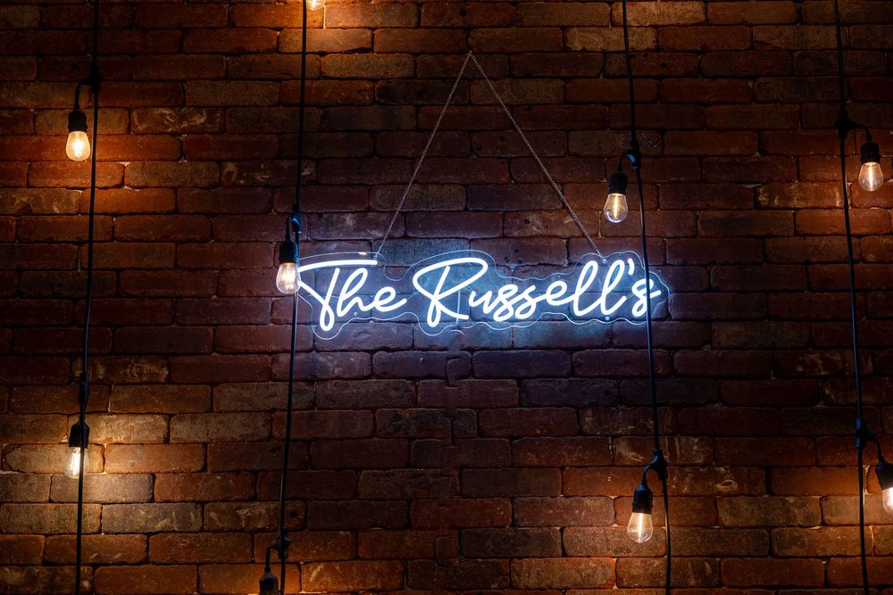 Rustic brick wall with hanging pendant lights and blue neon sign reading 'The Russell's' creates cozy ambiance.