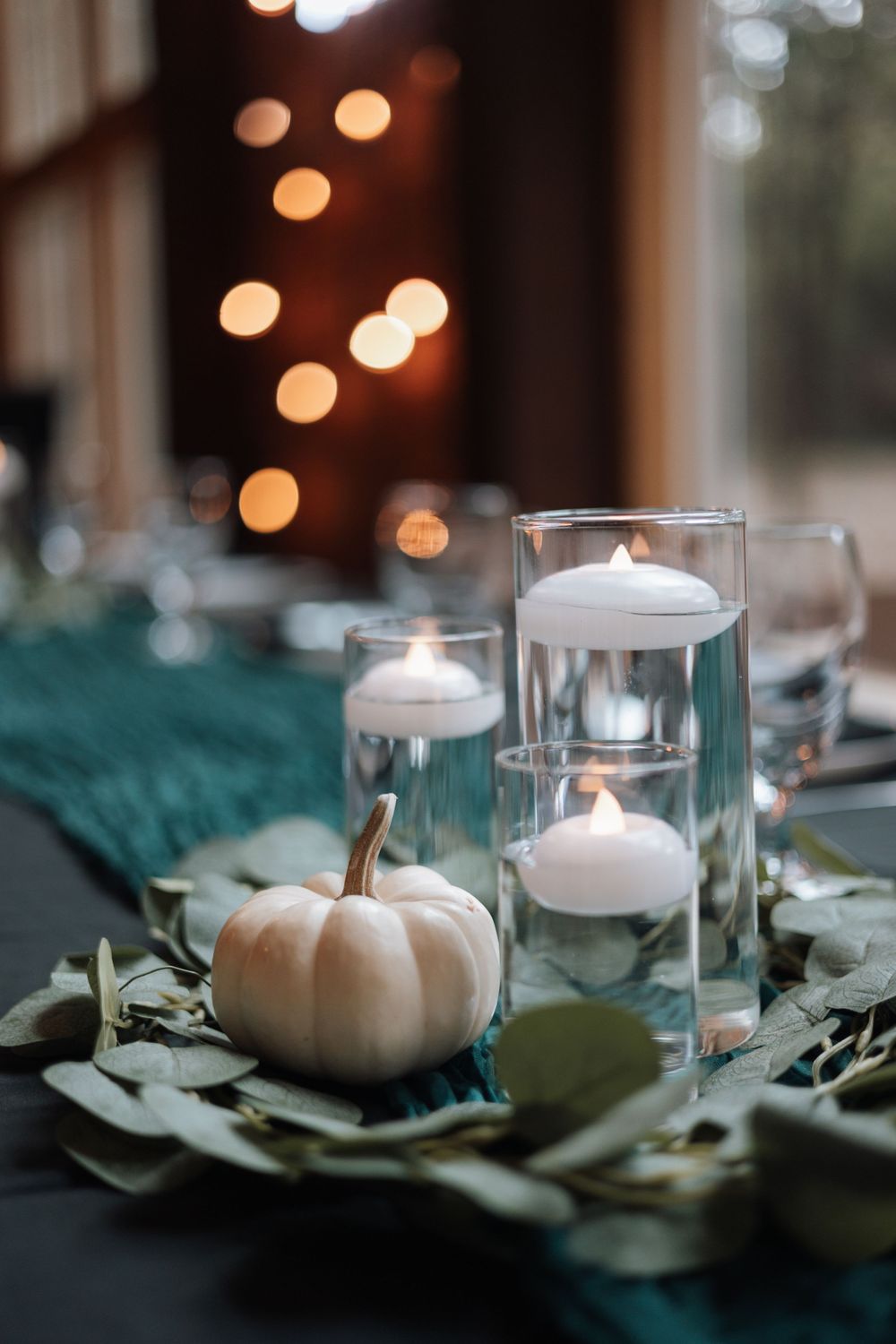 White candles and small pumpkins create an elegant autumn tablescape with soft bokeh lights in the background.