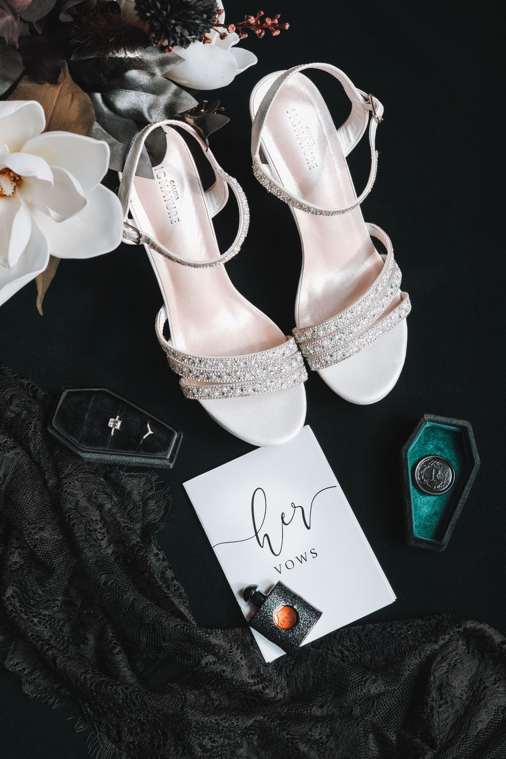 White wedding sandals with rhinestone straps displayed with nail polish and a handwritten note on black fabric.