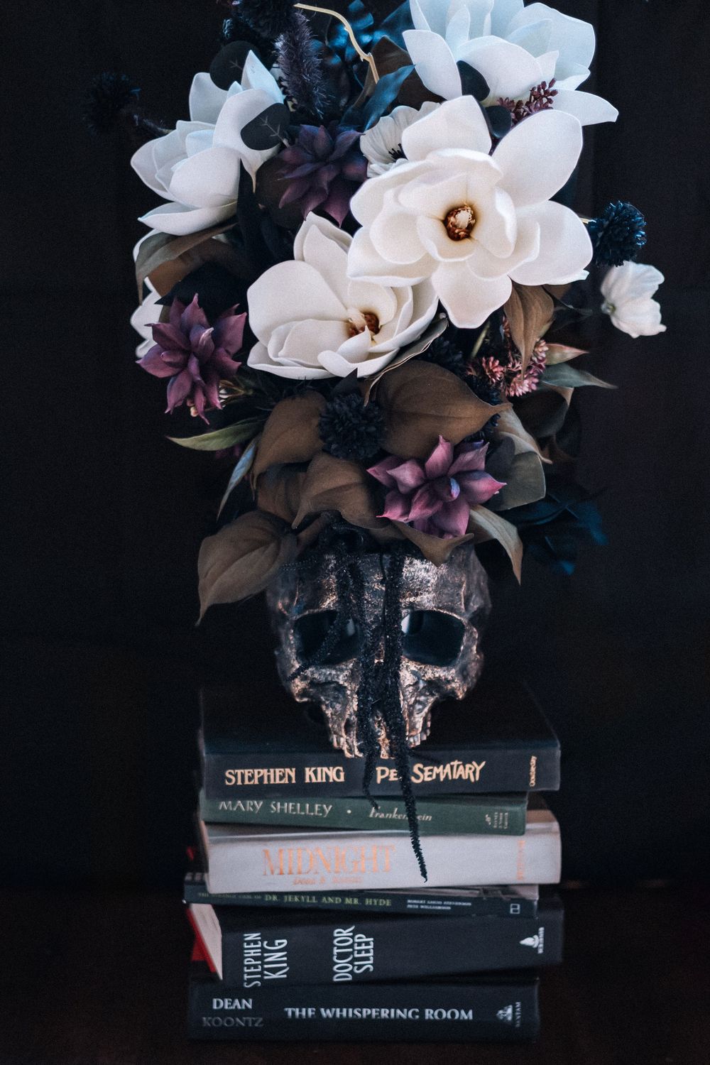 Dark moody still life with white magnolias and purple flowers in skull vase atop stack of Stephen King books.