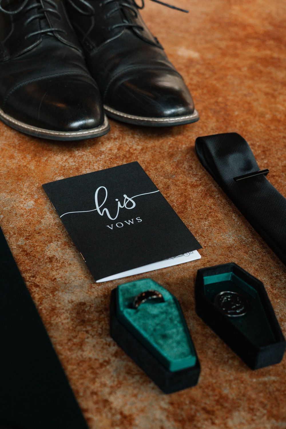 Black dress shoes and socks arranged next to a small black notebook and emerald velvet ring box on brown textured surface.