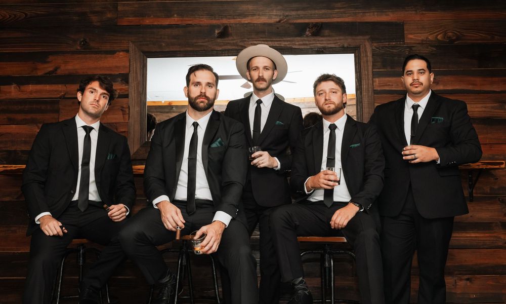 A group of groomsmen in black suits pose together against a rustic wooden wall background.