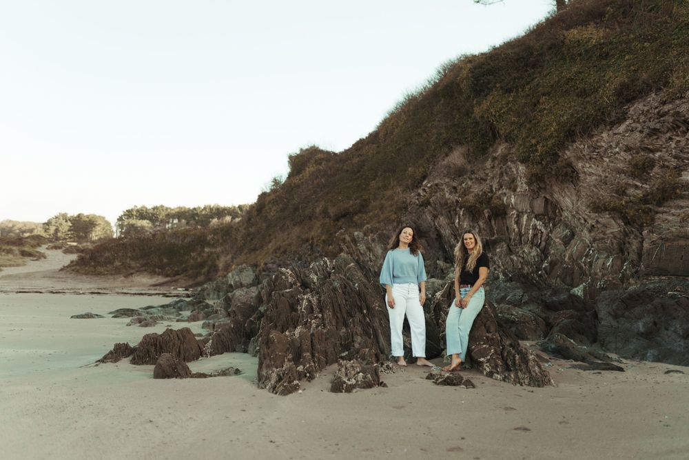 Two people stand on rocky coastal cliffs at sunset against a scenic beach backdrop.