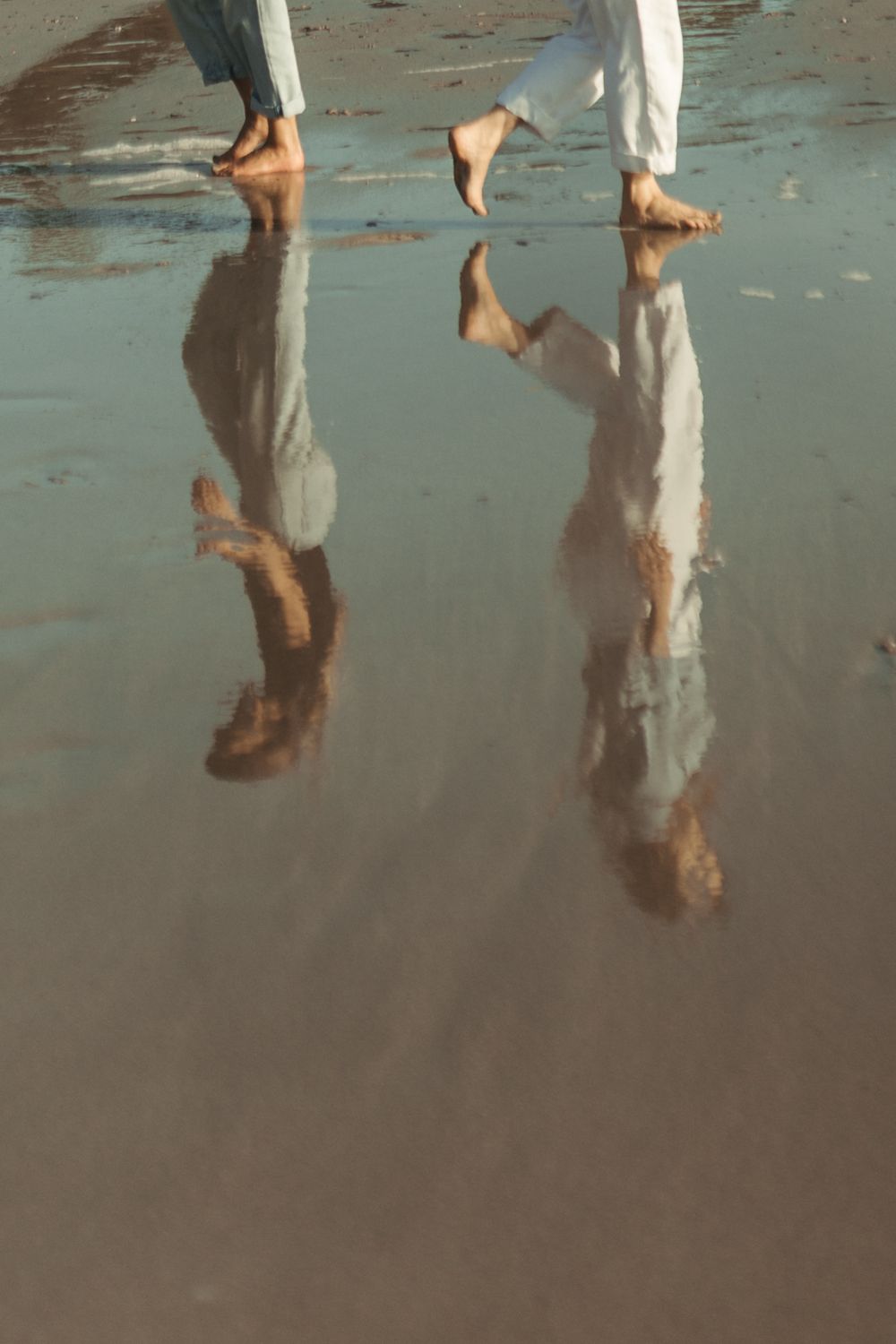 Reflections of feet and legs on wet sand at the beach during low tide create mirror-like symmetry in the water.
