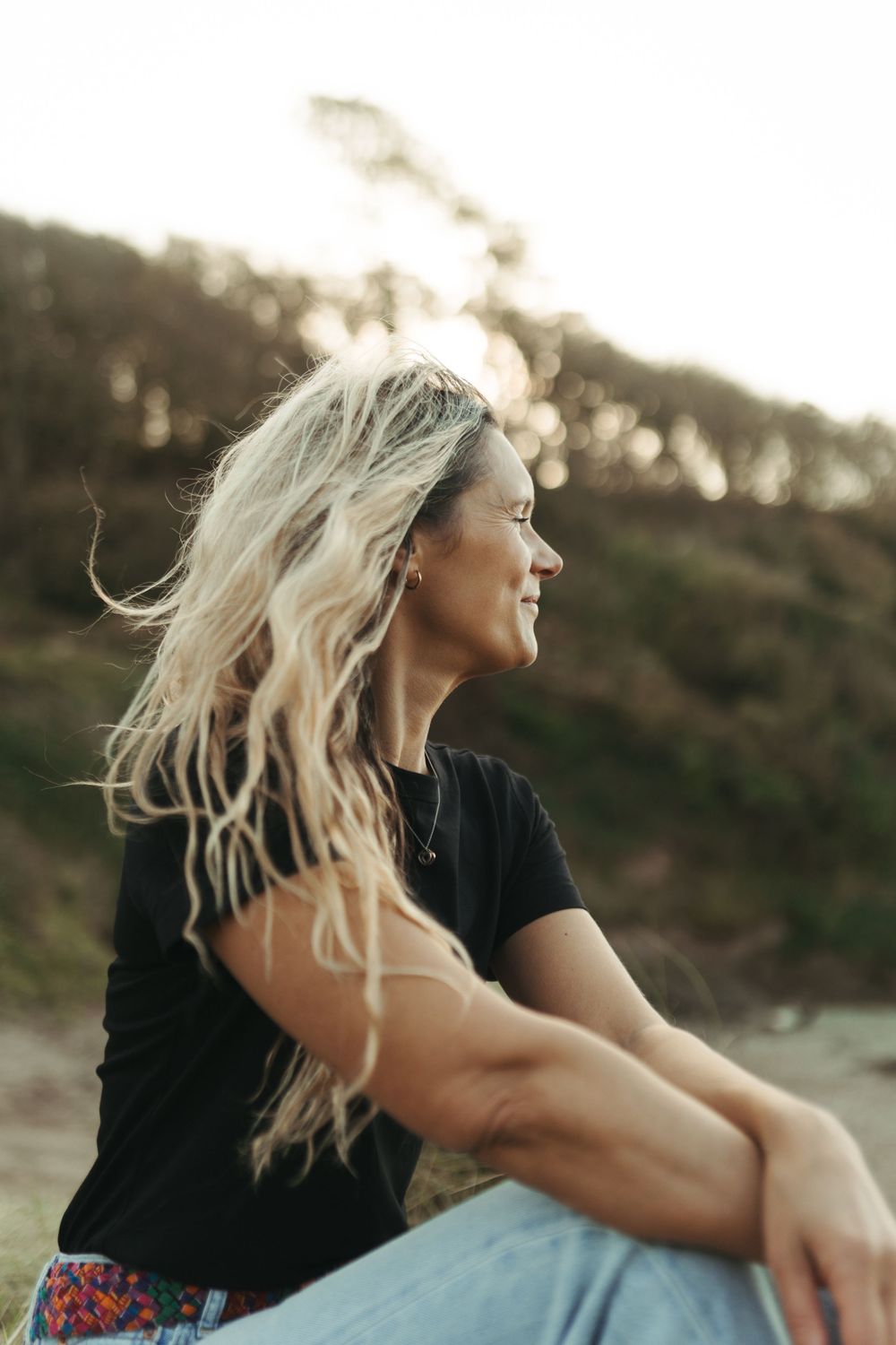 A person with flowing blonde hair laughs joyfully while sitting outdoors during golden hour.