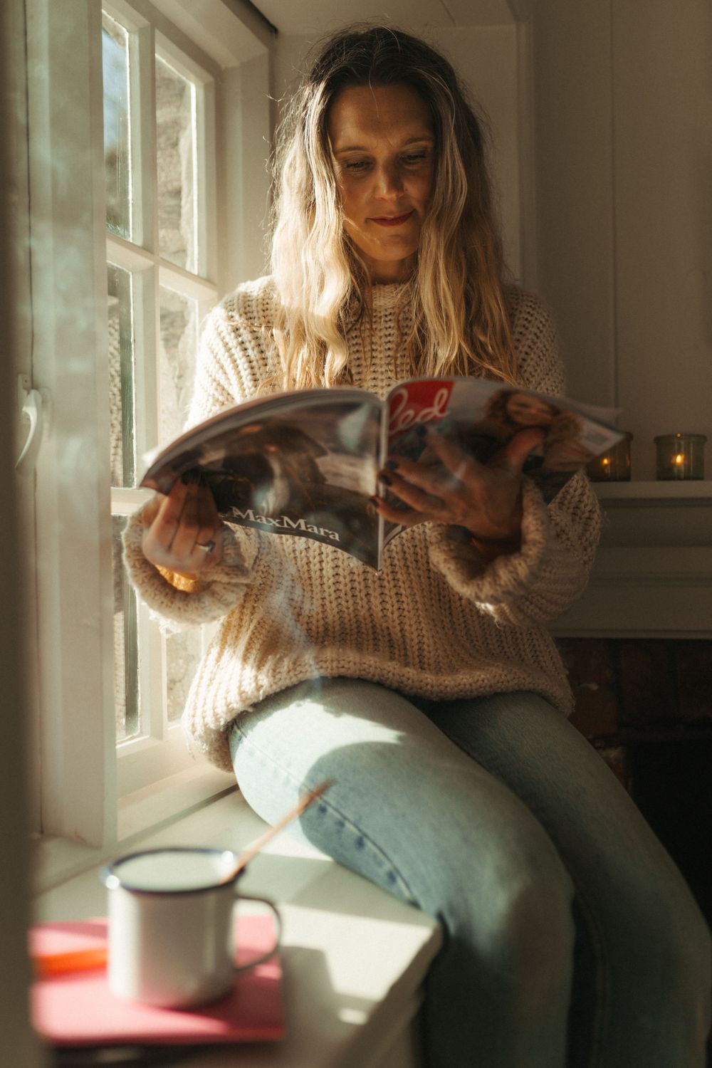 A person in a beige sweater sits by a window reading a book while natural light streams in through the glass.