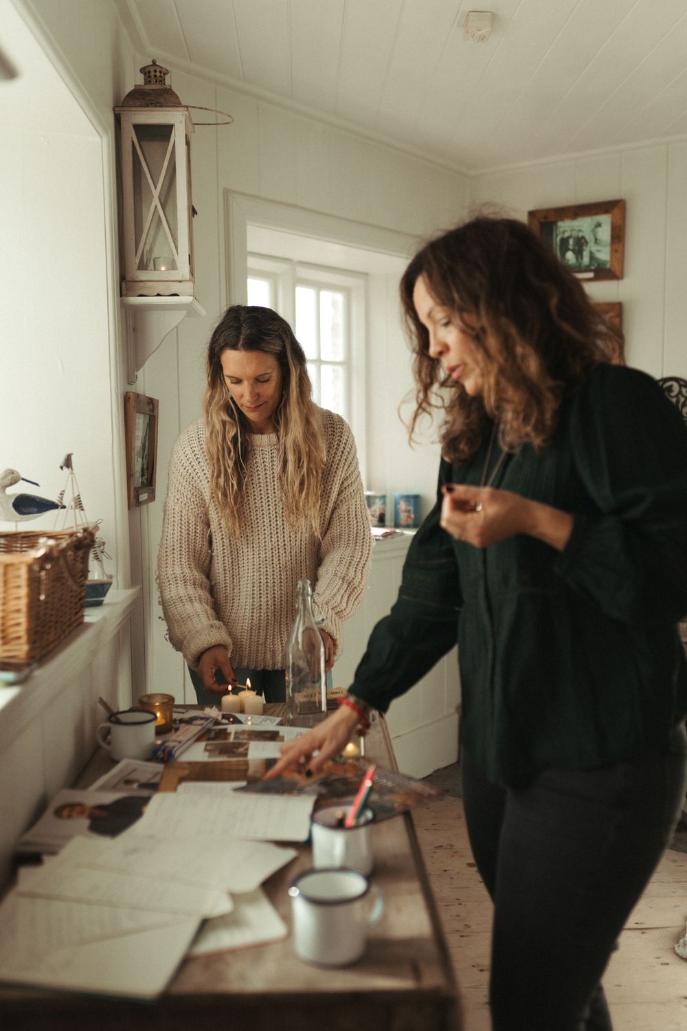 Two friends working together at a rustic wooden table in a bright cottage interior.