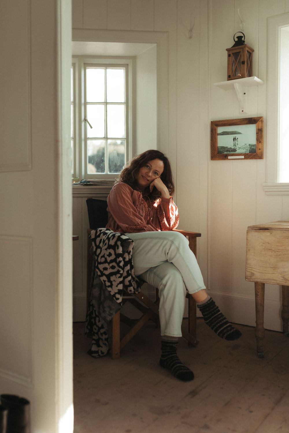 Moody interior scene of a white room with windows casting natural light and someone resting on a wooden chair.