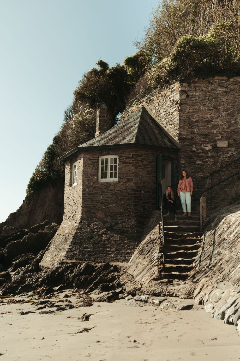A rustic stone cottage built into a rocky coastal cliff with steps leading down to a sandy beach at low tide.
