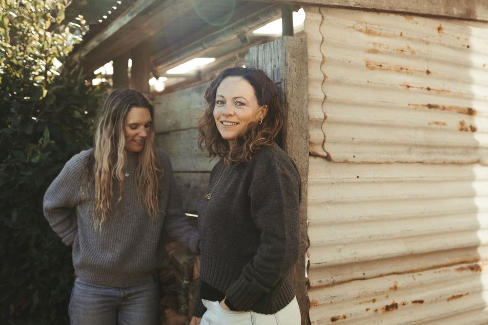 Two people in casual hoodies stand together outside a rustic wooden building on a sunny day.