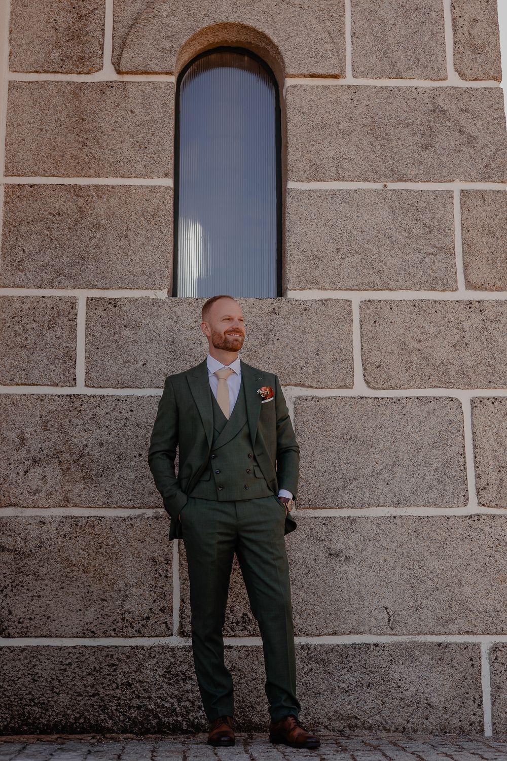 A groom in a dark green suit stands confidently against a stone wall with an arched window.