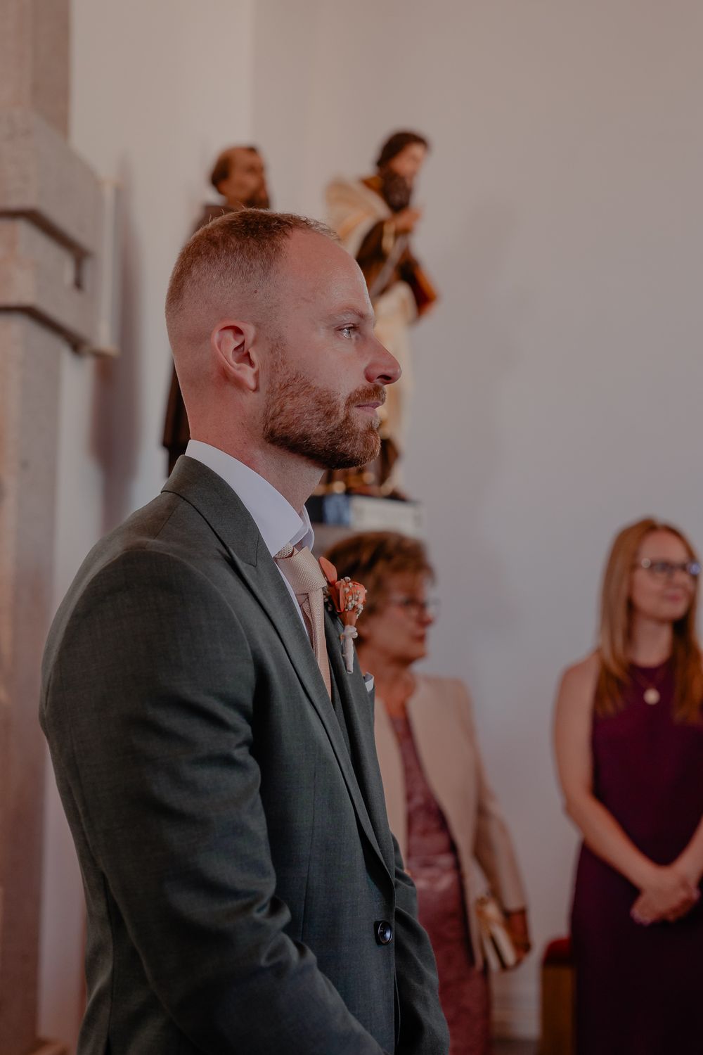 Wedding ceremony in progress inside church with decorative statues and burgundy-dressed attendees.
