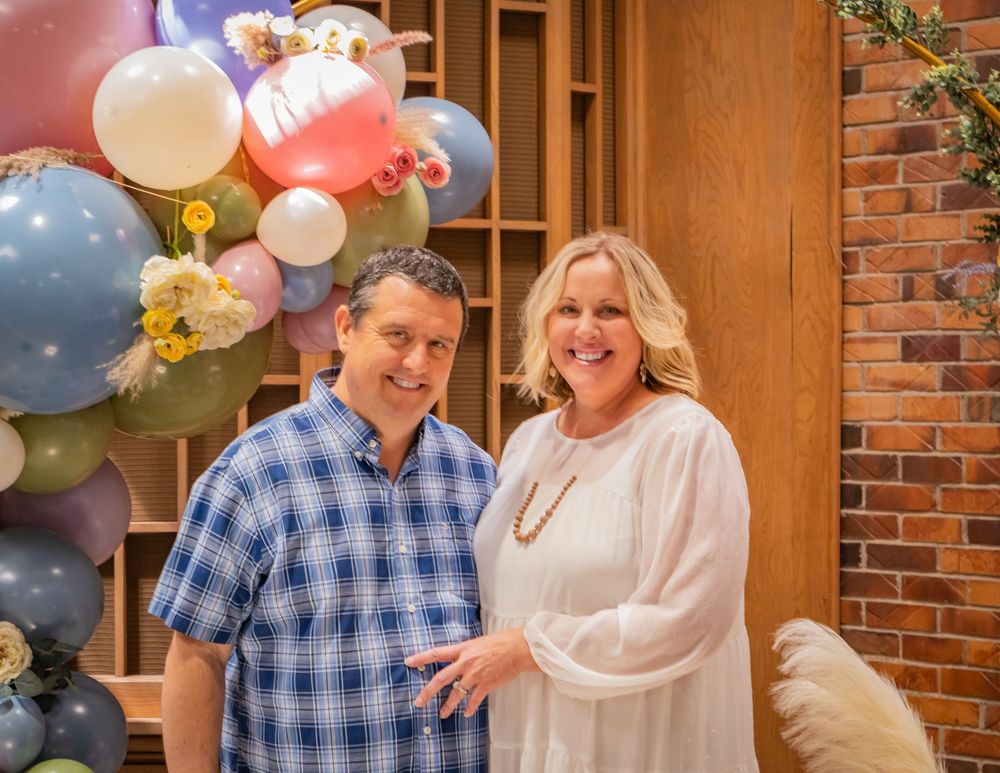 A couple celebrates at a party with colorful balloons and brick walls in the background.
