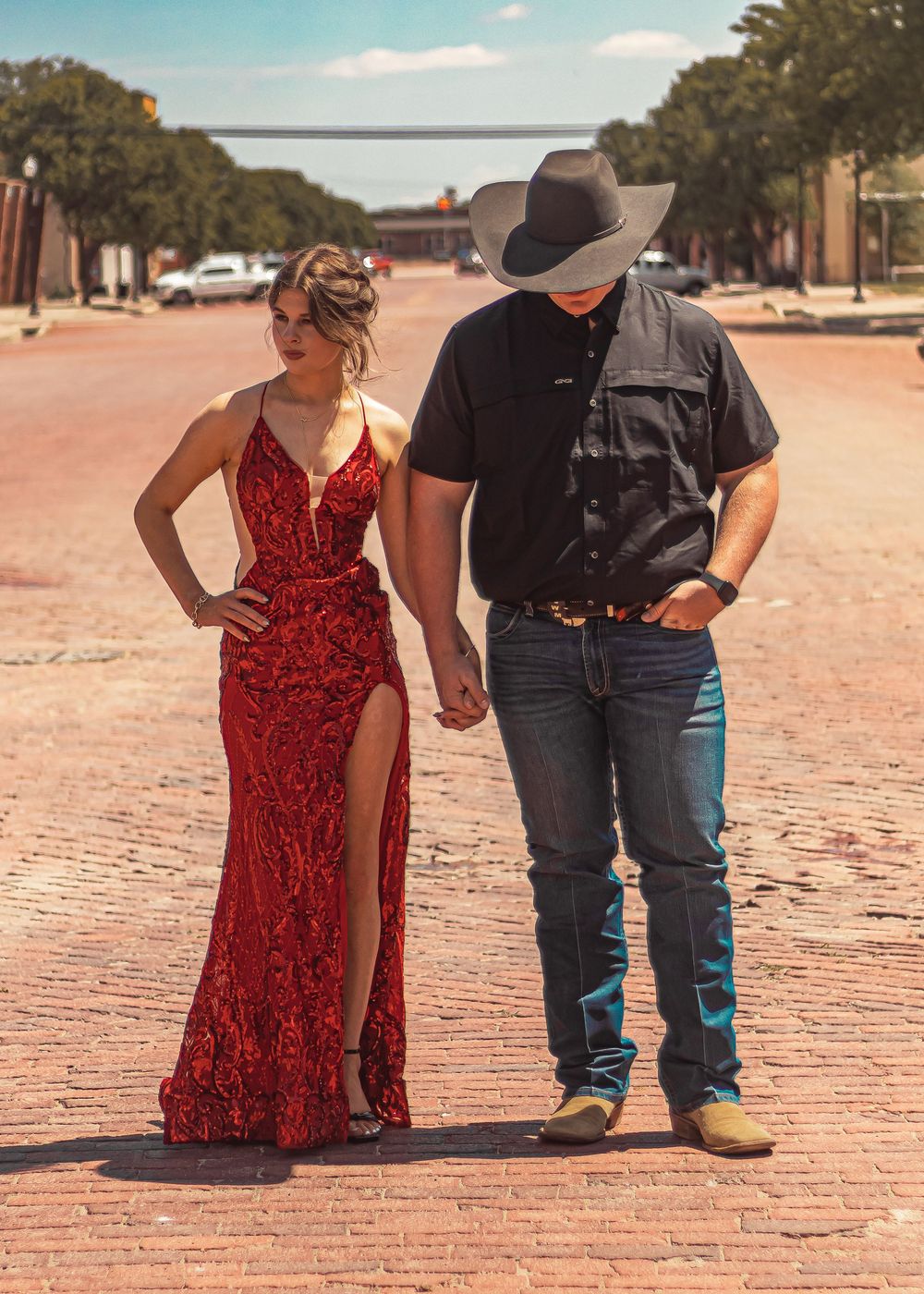 A couple in western attire, with a red formal dress and cowboy hat, holding hands on a brick street in a small town.