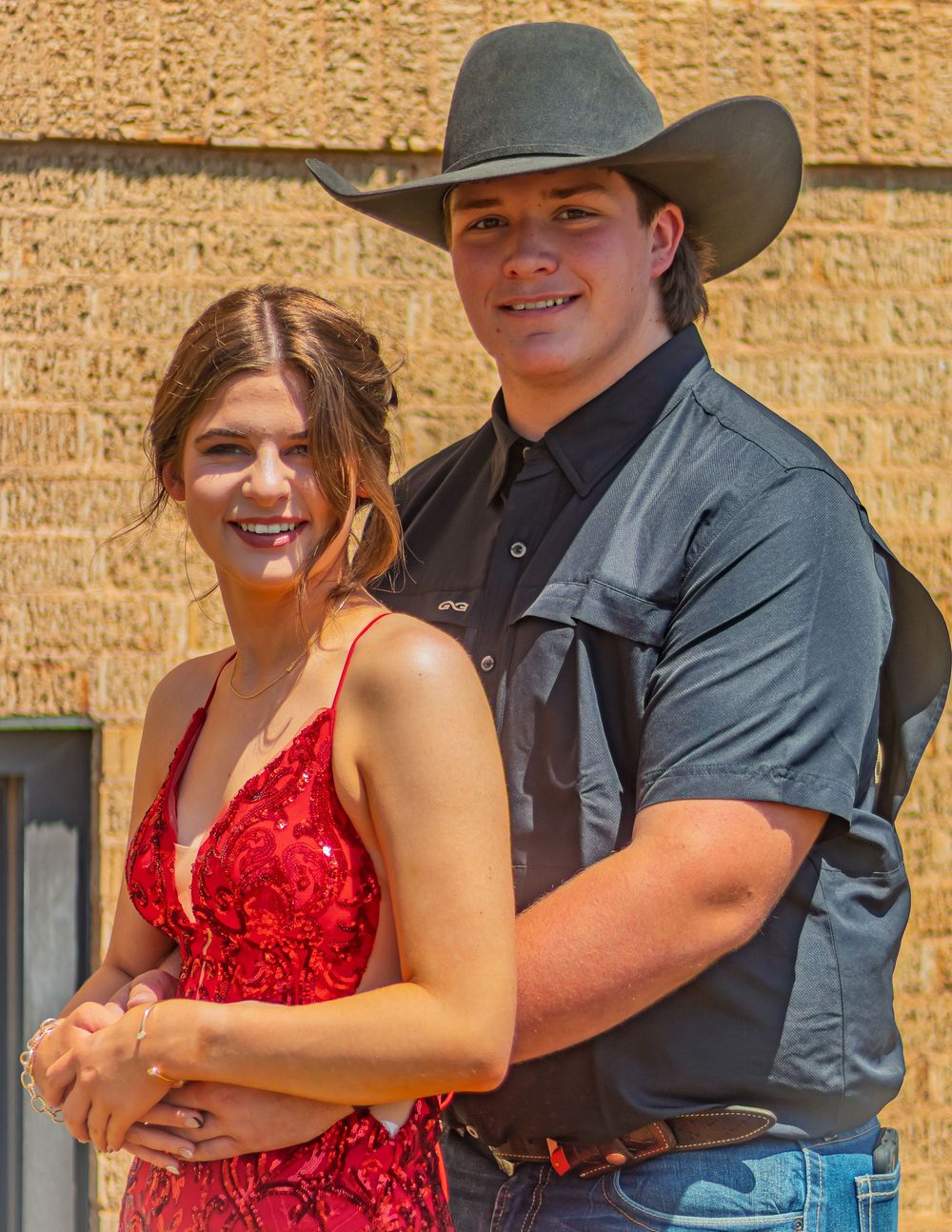 A couple poses together at a formal event, with one wearing a red dress and the other in western attire with a cowboy hat.