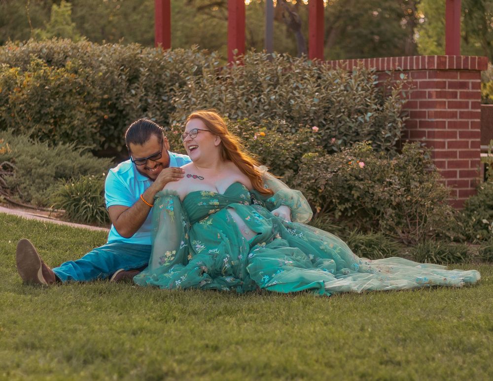 A couple in teal outfits relaxes together on a blanket in a grassy outdoor setting near a brick wall.
