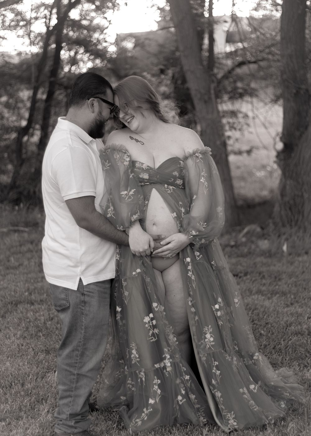 Black and white maternity photo showing couple in intimate pose against natural outdoor backdrop.