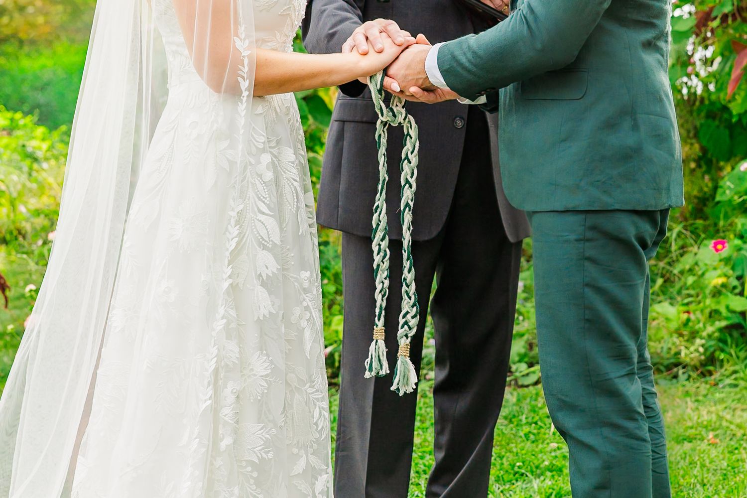 A couple in a green outdoor setting during their wedding ceremony with flowing white dress details.