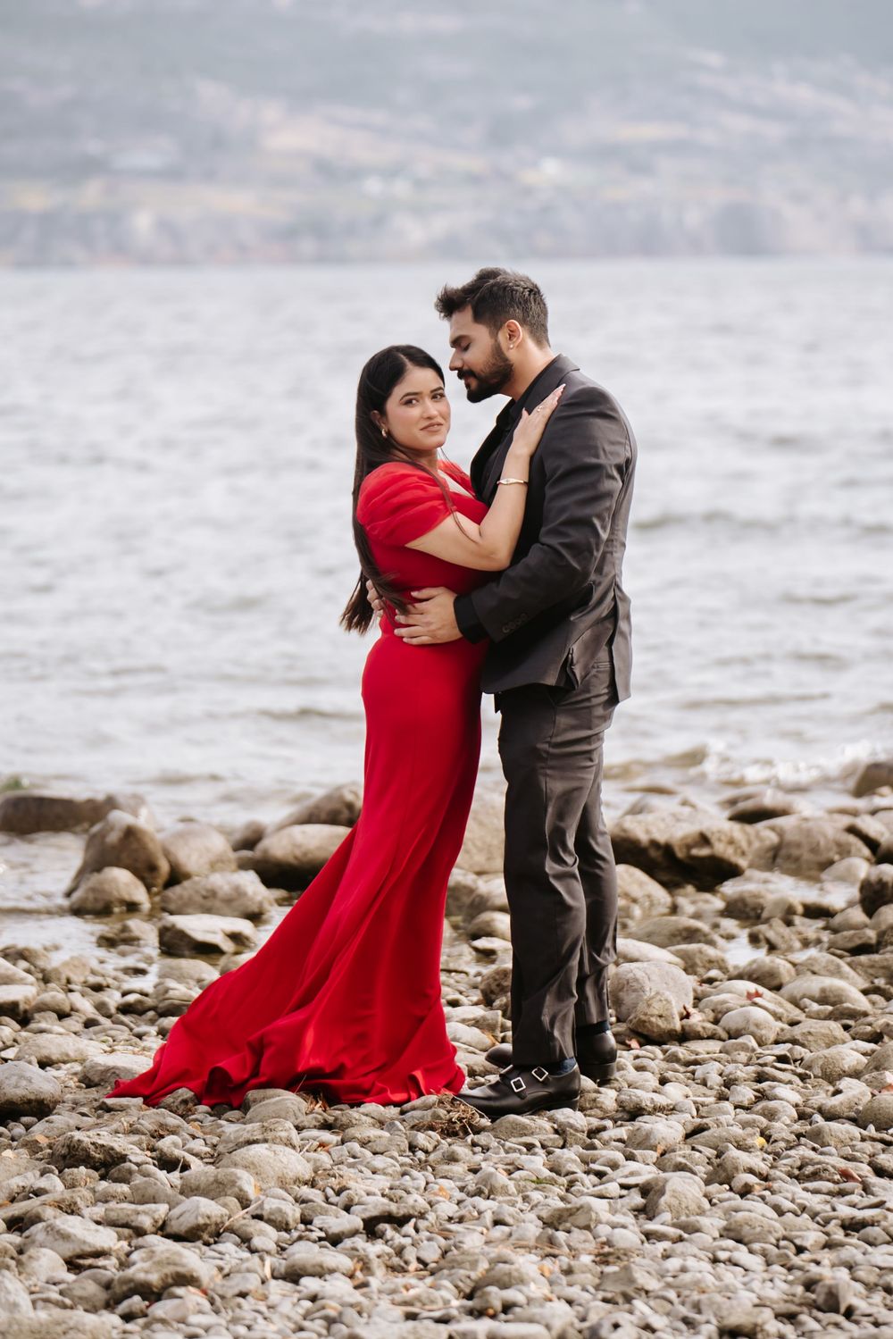 A couple in formal attire embraces romantically on a rocky beach shore as waves crash behind them.