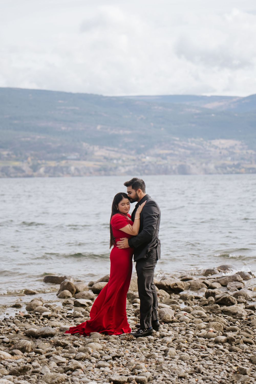 A couple embraces on a rocky beach shore with mountains and lake in the background.