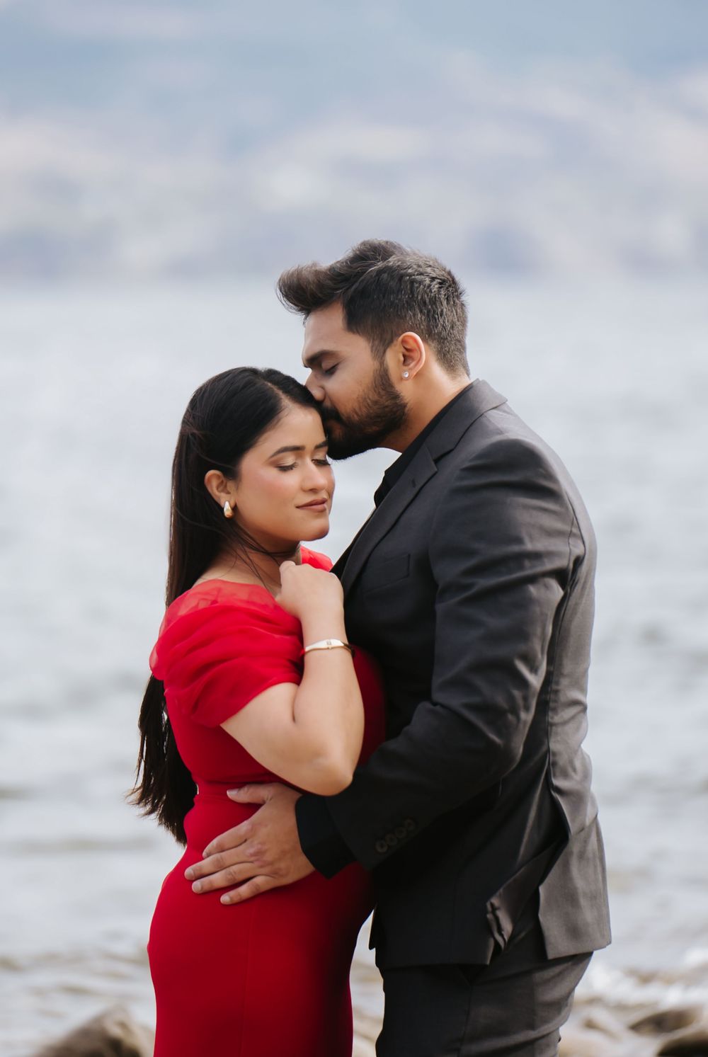 A couple in red dress and black suit share intimate moments during a romantic photoshoot by the ocean.