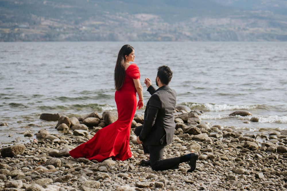 Two people sit together on a rocky beach overlooking a scenic mountain lake on a cloudy day.