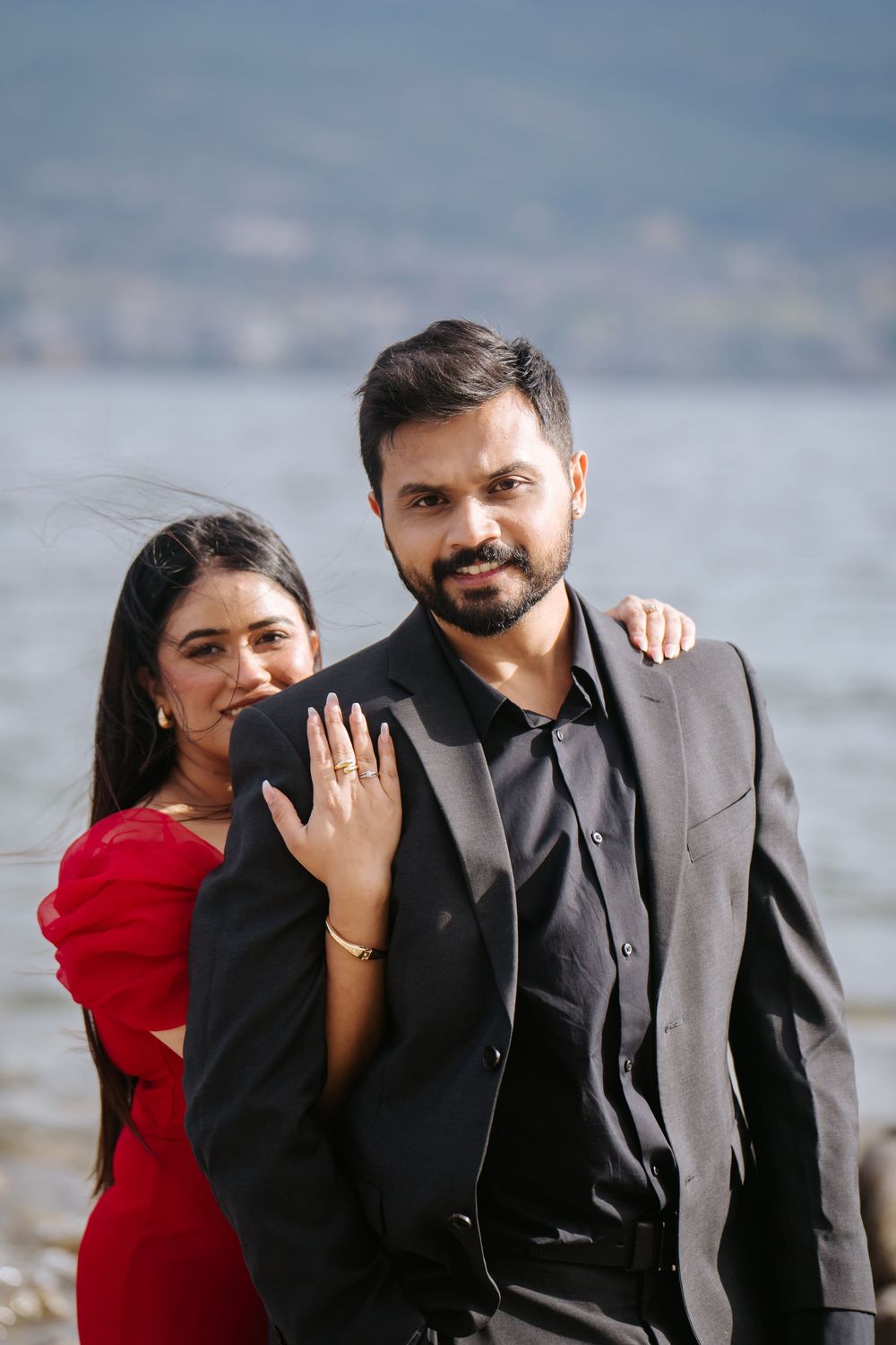 A couple poses together by the water, with the woman in a red dress showing off her engagement ring.