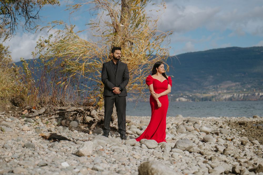A couple in formal attire poses together on a rocky beach with mountains and autumn trees in the background.