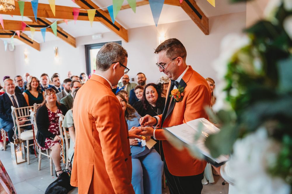 Gay couple in orange suits Indoor celebration venue with exposed beams and colorful bunting decorations. Gellifawr Woodland Retreat