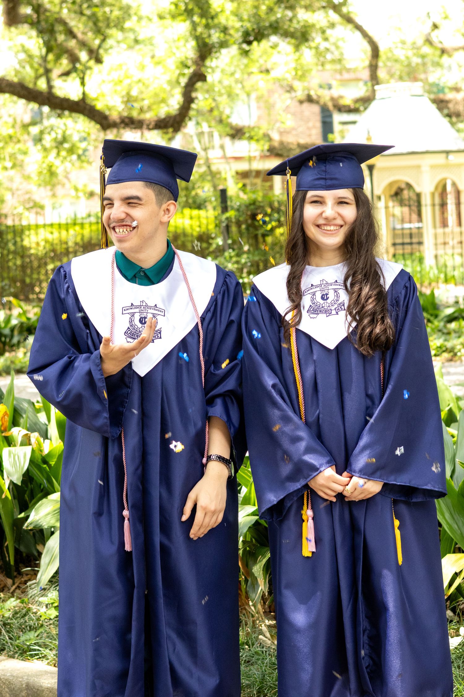 Two graduates in navy blue caps and gowns stand together smiling outdoors on a sunny day.